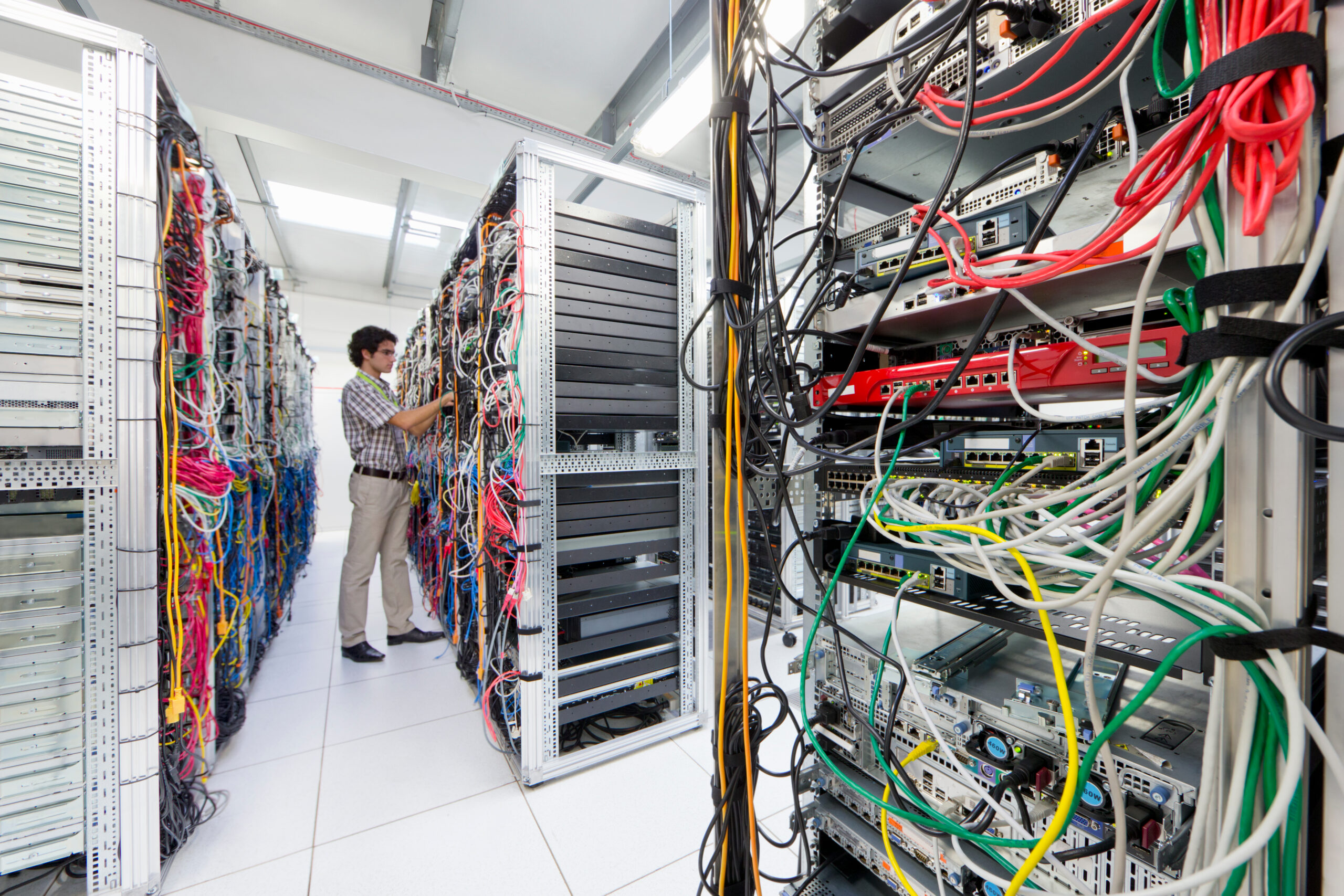 Wide shot inside a commercial data centre aisle with a technician checking cable terminations on a densely-populated server rack